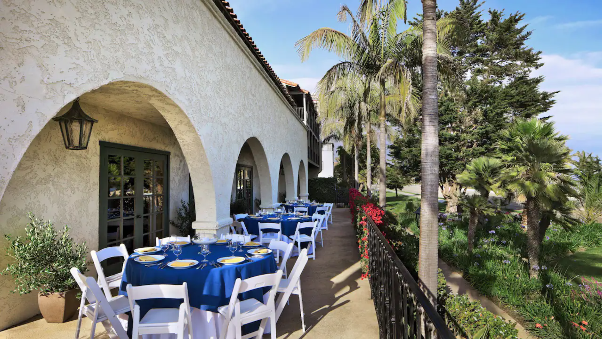 Outdoor seating area with rounded tables, blue linens and white chairs near palm trees at Mar Monte Hotel in Santa Barbara, CA