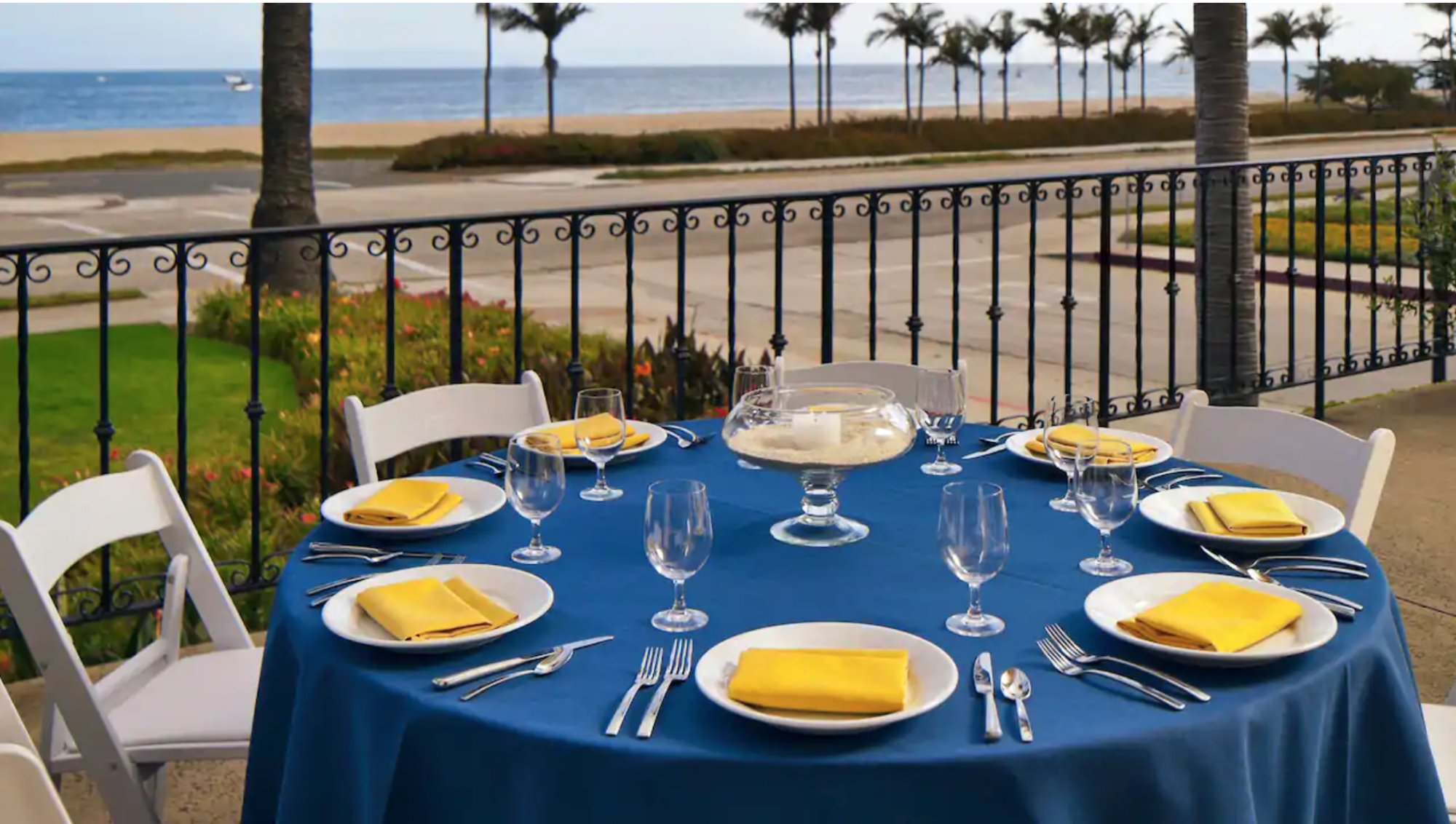 Round table with blue linens, yellow napkins, white plates and glassware overlooking roadway, beach and palm trees at Mar Monte Hotel in Santa Barbara, CA