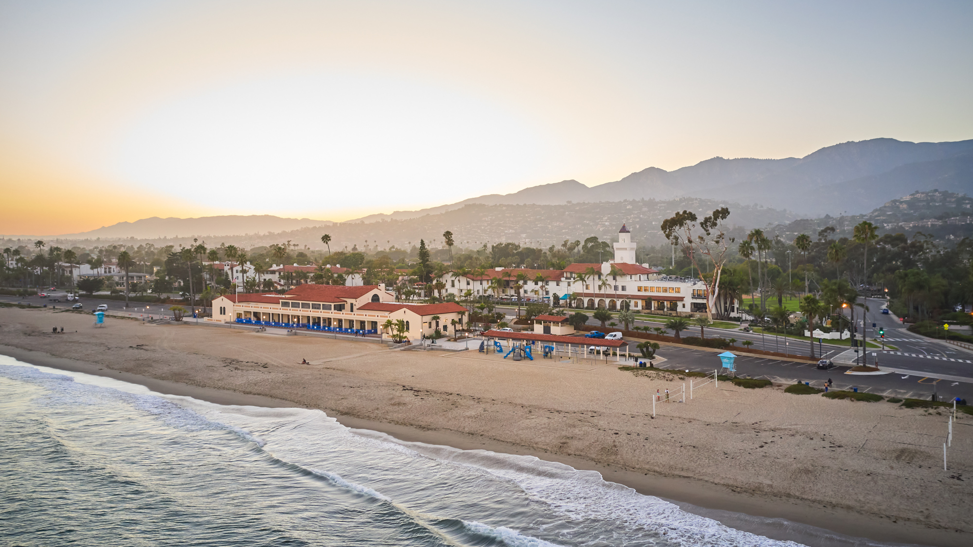 Aerial view of sandy beach at sunset with people on it near ocean with Mar Monte Hotel in Santa Barbara, CA behind the beach with mountains in the background