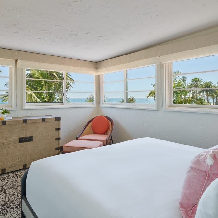 Large bed with white linens and pink pillows near red chair overlooking ocean and palm trees at Mar Monte Hotel in Santa Barbara, CA