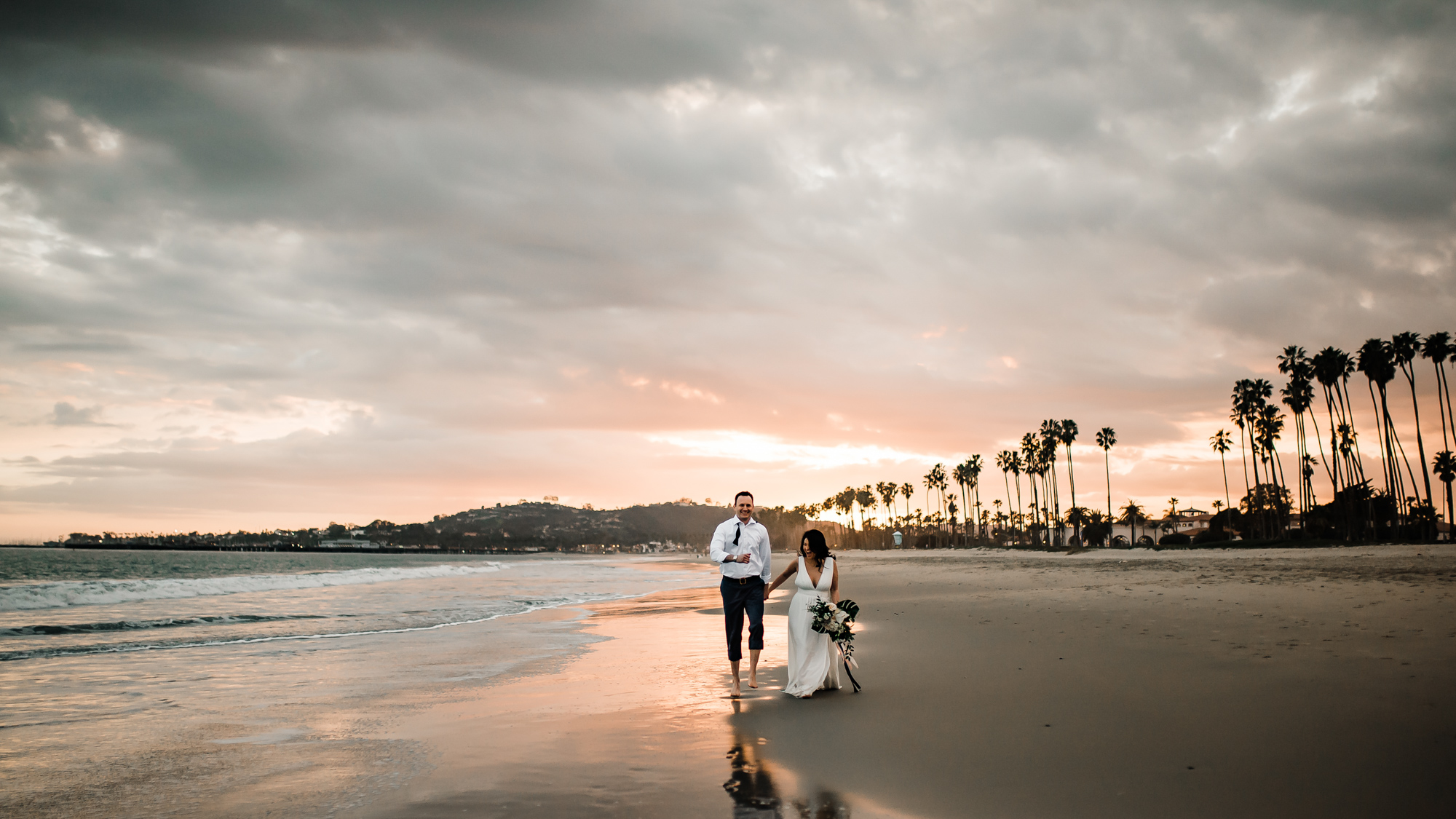 A couple holding hands walking on the beach with wedding bouquet at sunset near palm trees at Mar Monte Hotel in Santa Barbara, CA