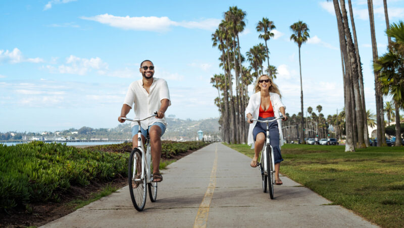 Guy and girl riding bicycles wearing swimsuits and sunglasses, smiling and laughing with each other