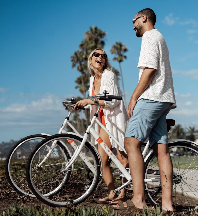 Guy and girl riding bicycles wearing swimsuits and sunglasses, smiling and laughing with each other