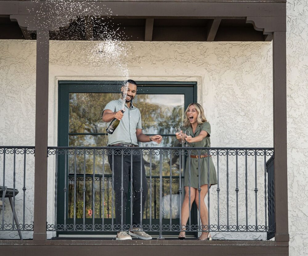 An attractive couple smiling and laughing that popped a bottle of champagne on guest room balcony