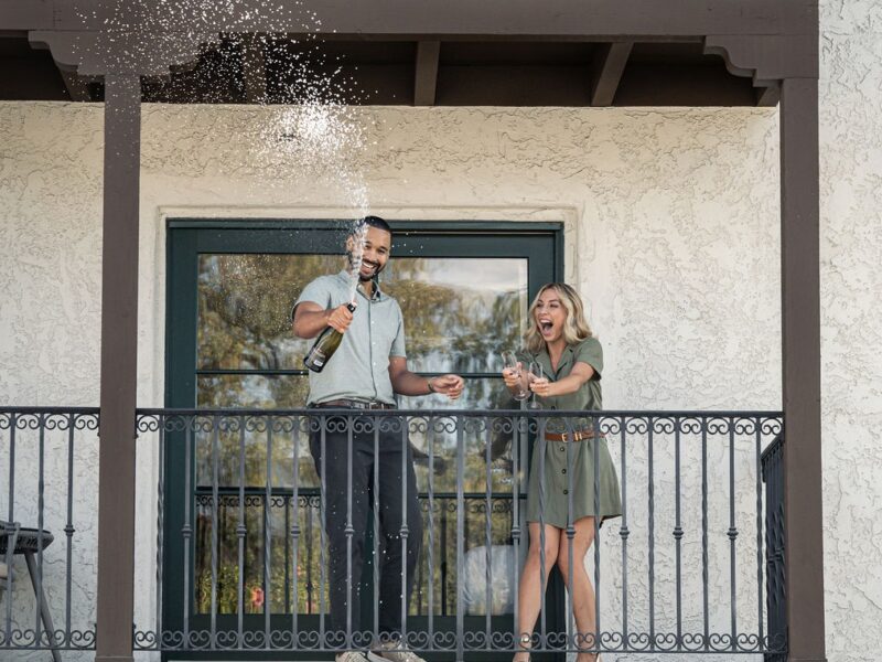 An attractive couple smiling and laughing that popped a bottle of champagne on guest room balcony