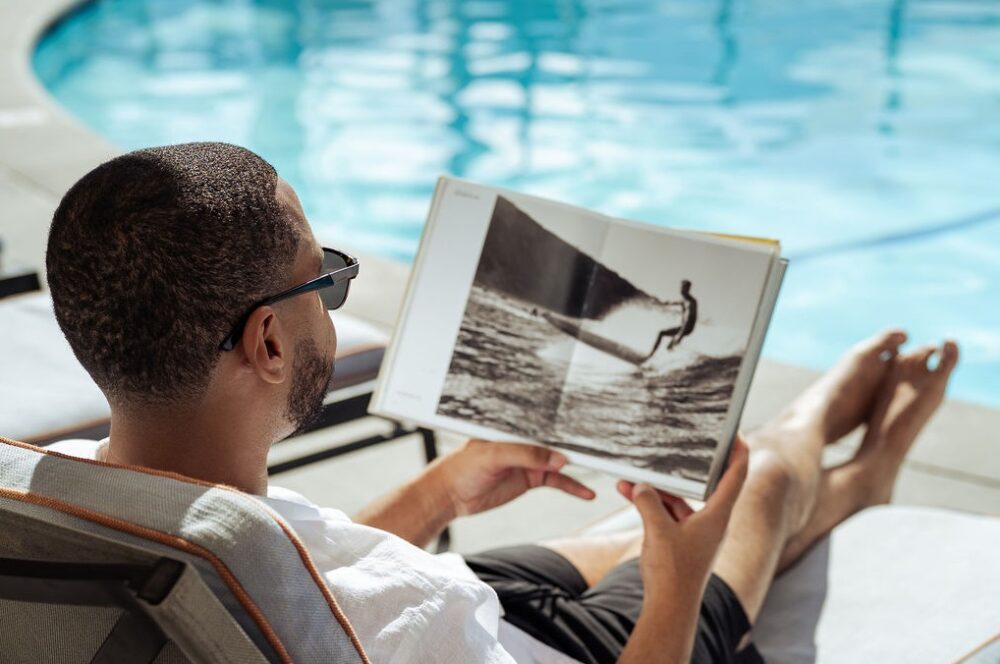 Man wearing sunglasses sitting poolside reading a surf book