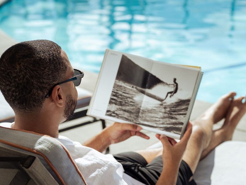 Man wearing sunglasses sitting poolside reading a surf book