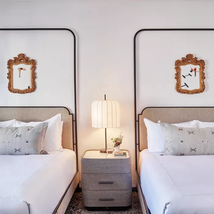 Two beds with white linens and grey pillows near side table with lamp below two clocks at Mar Monte Hotel in Santa Barbara, CA